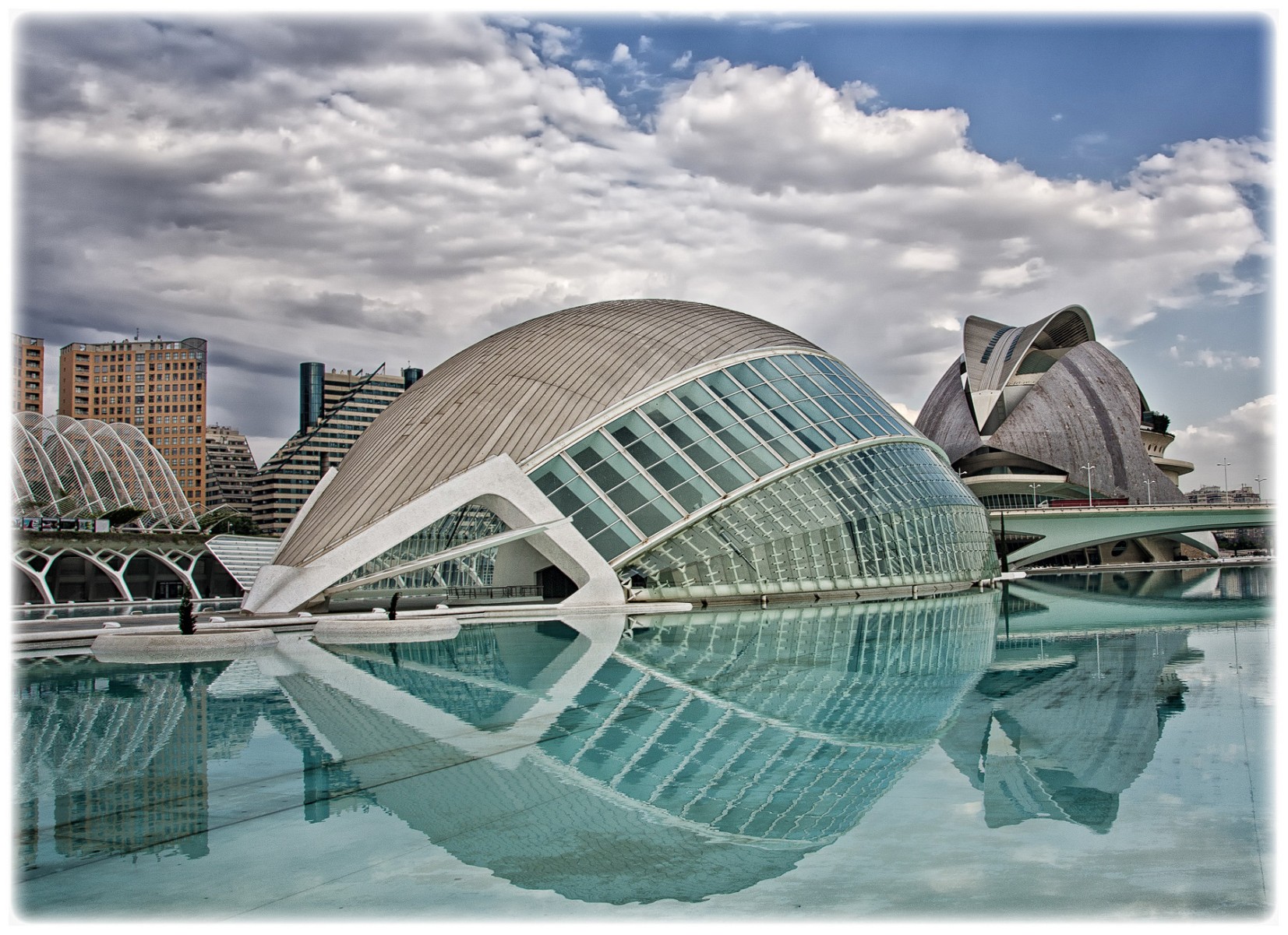 Futuristische, kuppelförmige Glas- und Stahlkonstruktion der Stadt der Künste und der Wissenschaften in Valencia mit Spiegelung im Wasser und bewölktem Himmel
